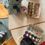 Zwei Kinder malen mit Farben auf einem Tisch, umgeben von leeren Eierkartons und Farbpaletten.